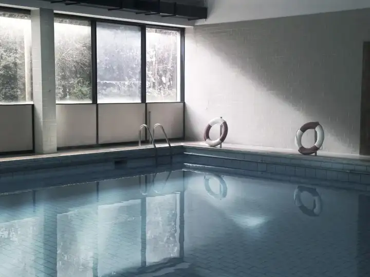 Leeres Hallenbad mit ruhigem Wasser, Rettungsringen an der Wand und großen Fenstern