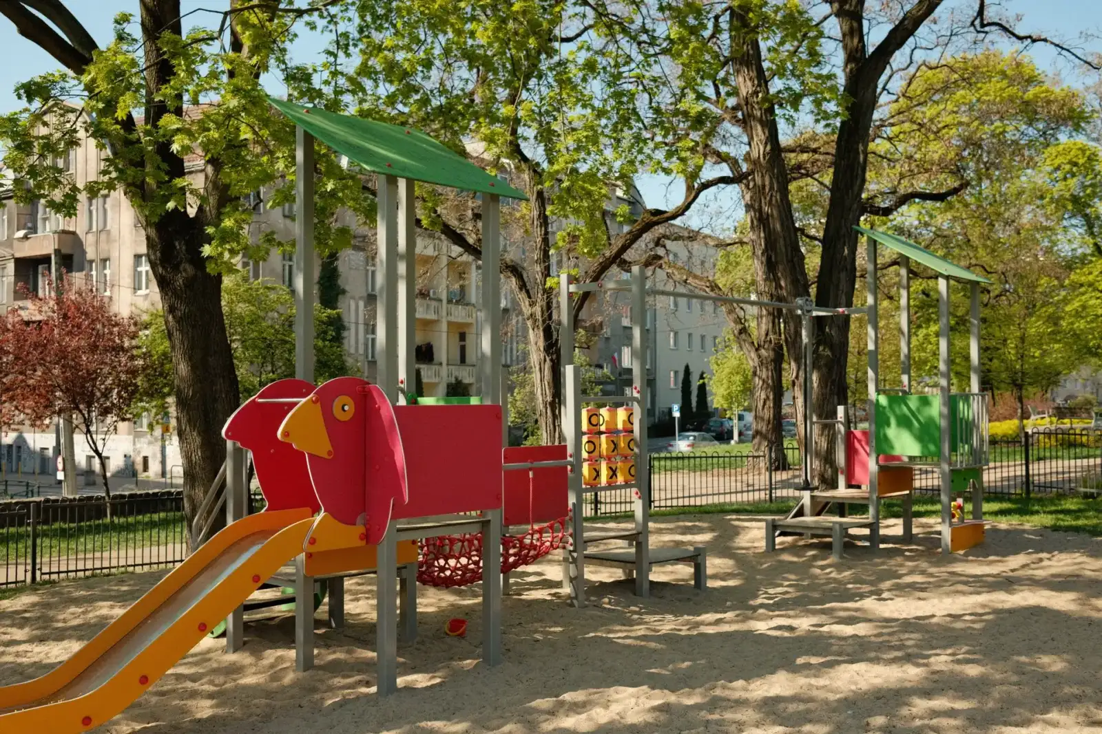 Spielplätze in meiner Nähe: Bunter Spielplatz mit Rutsche und Spielgeräten auf Sandboden zwischen hohen Bäumen