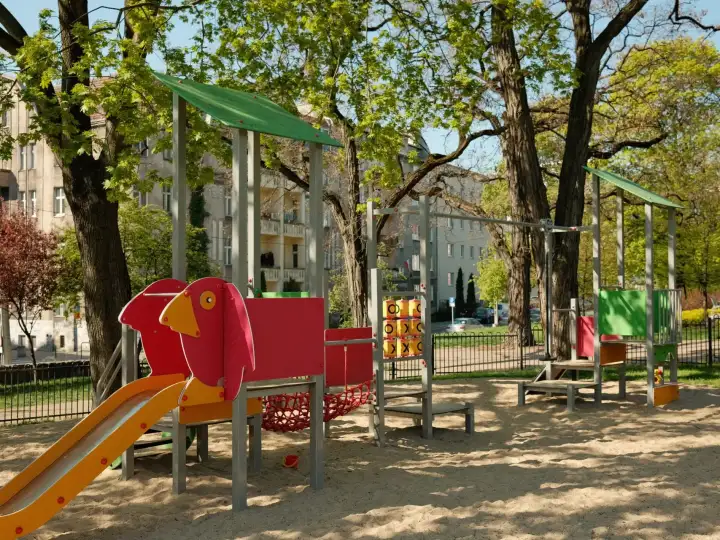 Spielplätze in meiner Nähe: Bunter Spielplatz mit Rutsche und Spielgeräten auf Sandboden zwischen hohen Bäumen