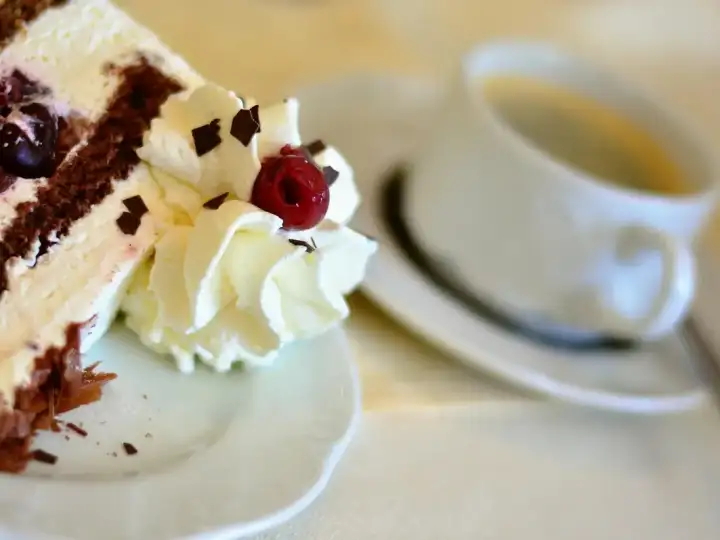 Stück Kirschtorte mit Sahne und Schokospänen neben einer Tasse Kaffee auf weißem Porzellan