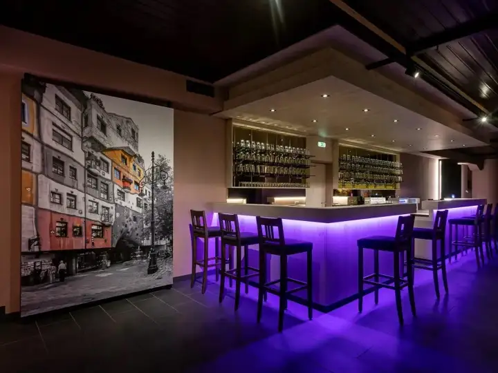 Violett beleuchtete Bar mit Barhockern, Glasregalen und Hundertwasserhaus-Wandbild